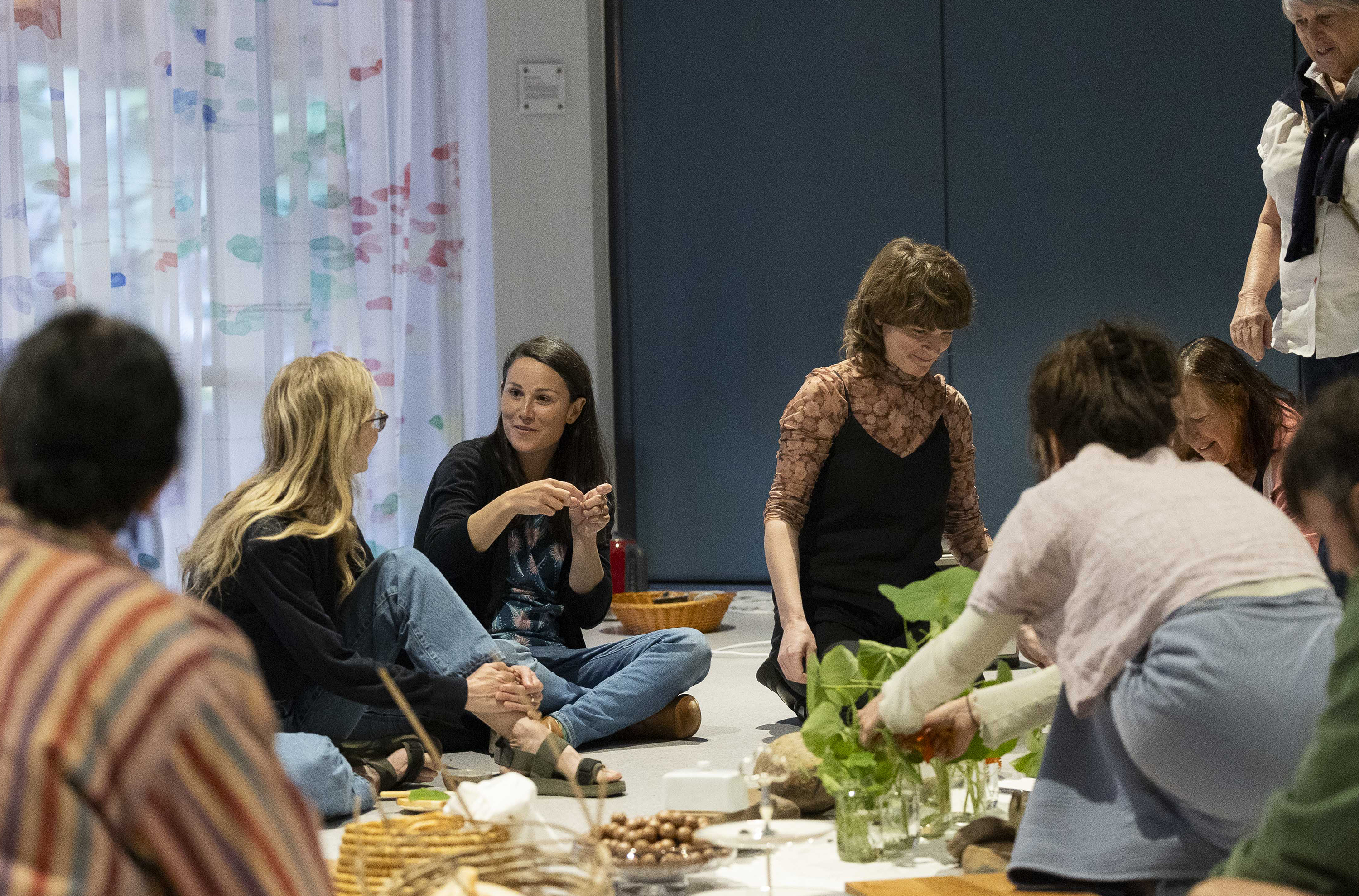 A group of people sitting on the floor and engaged in conversation around an informal tea setting including nasturtium plants and macadami nuts